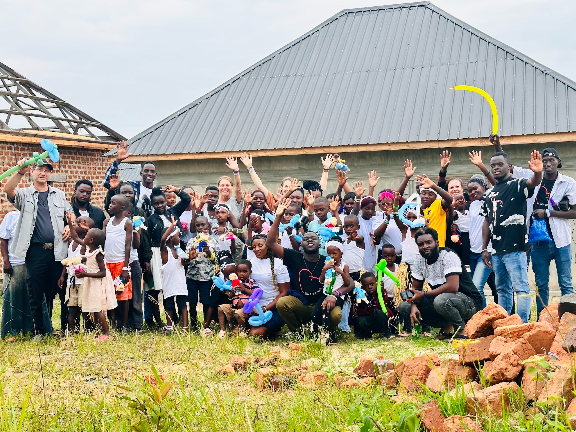 Photo of a large group of smiling and waving people in front of a small house