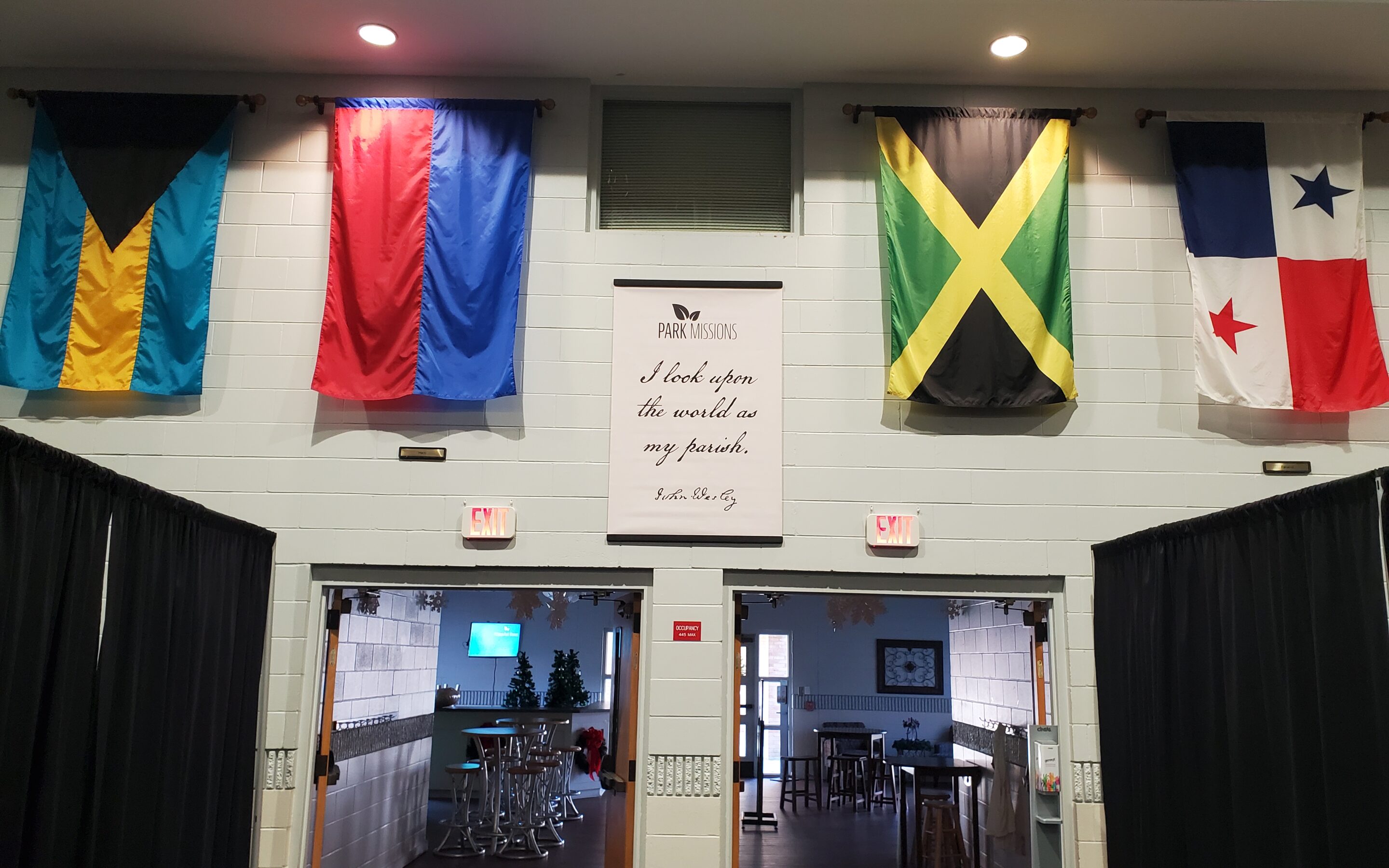 Photo of the wall above the Fellowship Hall exit doors with two flags on either side of a banner that says "Park Missions I look upon the World as my Parish -John Wesley"