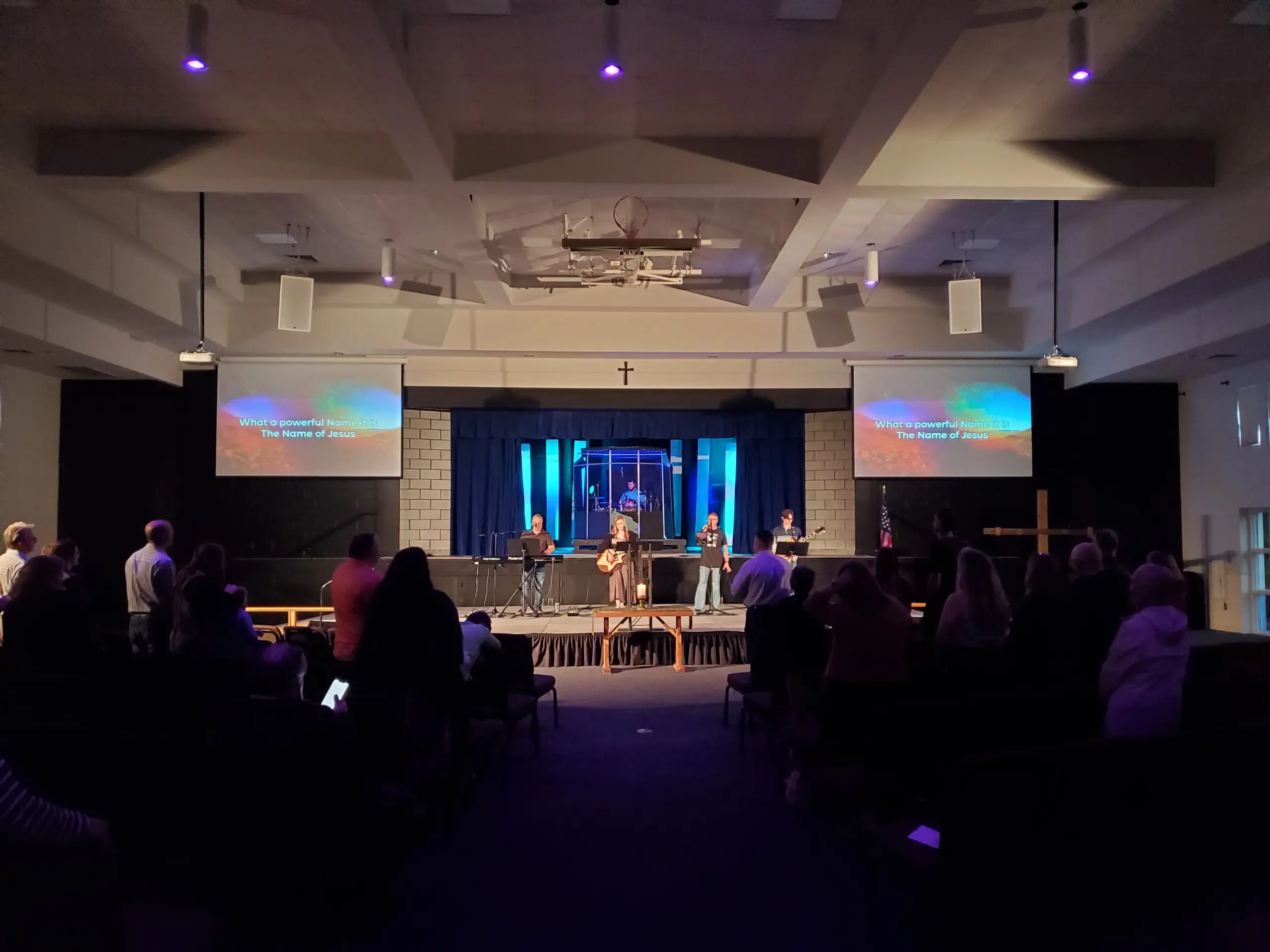 View of a lit stage from the back of a dark room with a crowd of people standing and facing the stage