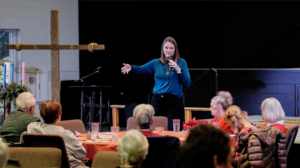 Women with microphone addresses a group of people sitting around a table in the Fellowship Hall.
